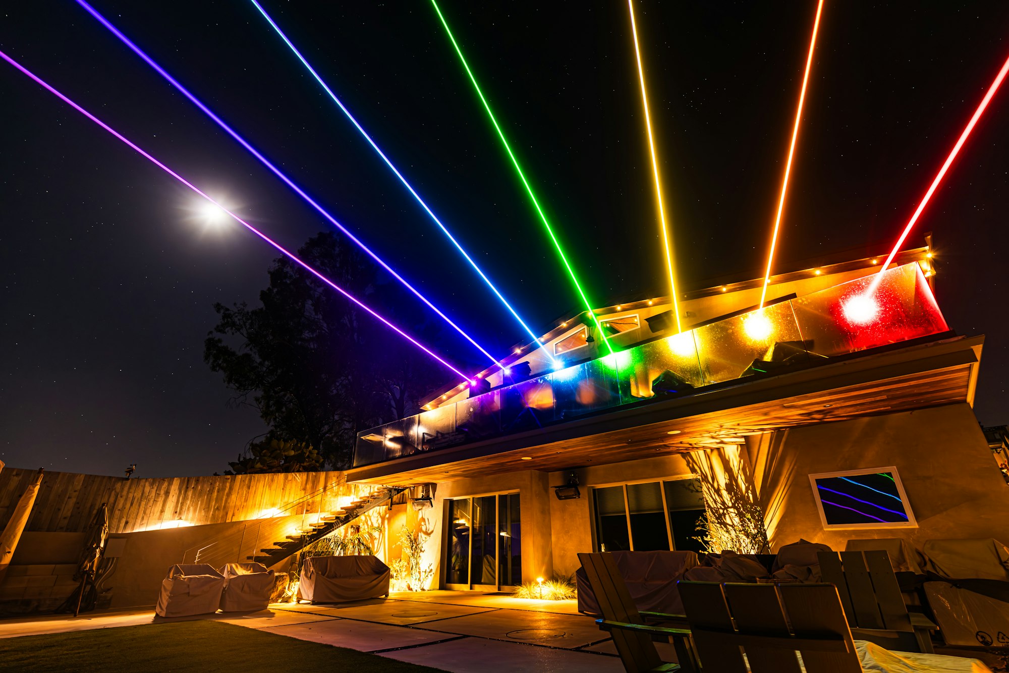 A stylish house illuminated by colorful laser beams against a night sky, with the moon glowing above and a serene outdoor space.