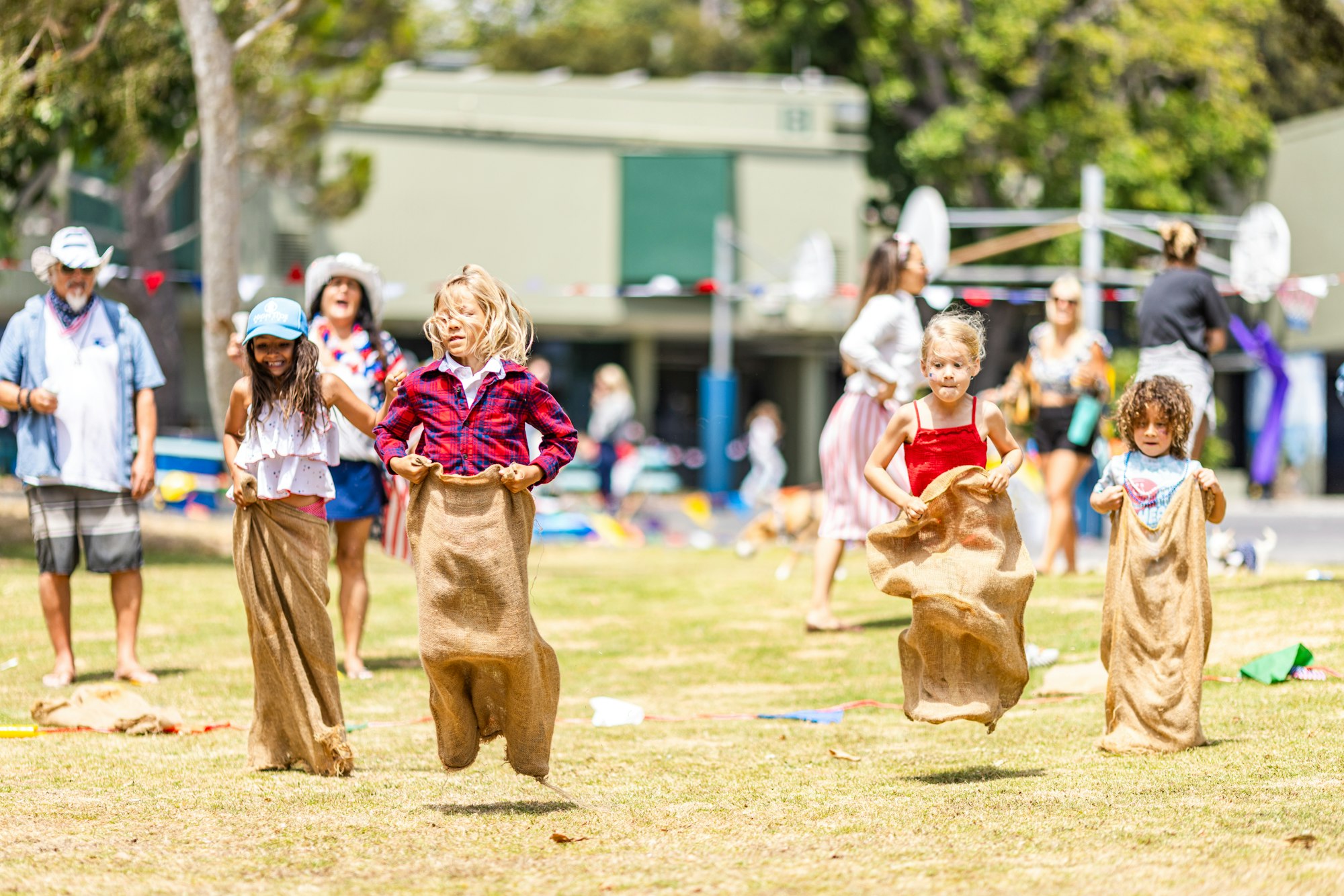 Children are participating in a sack race outdoors, with onlookers in the background.