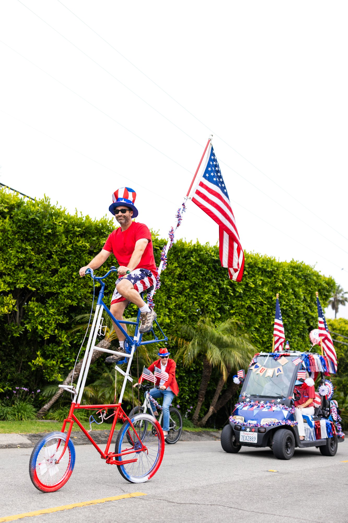 Man on tall bike wearing patriotic colors, holding a USA flag, in a festive parade with a decorated golf cart and others in similar attire.