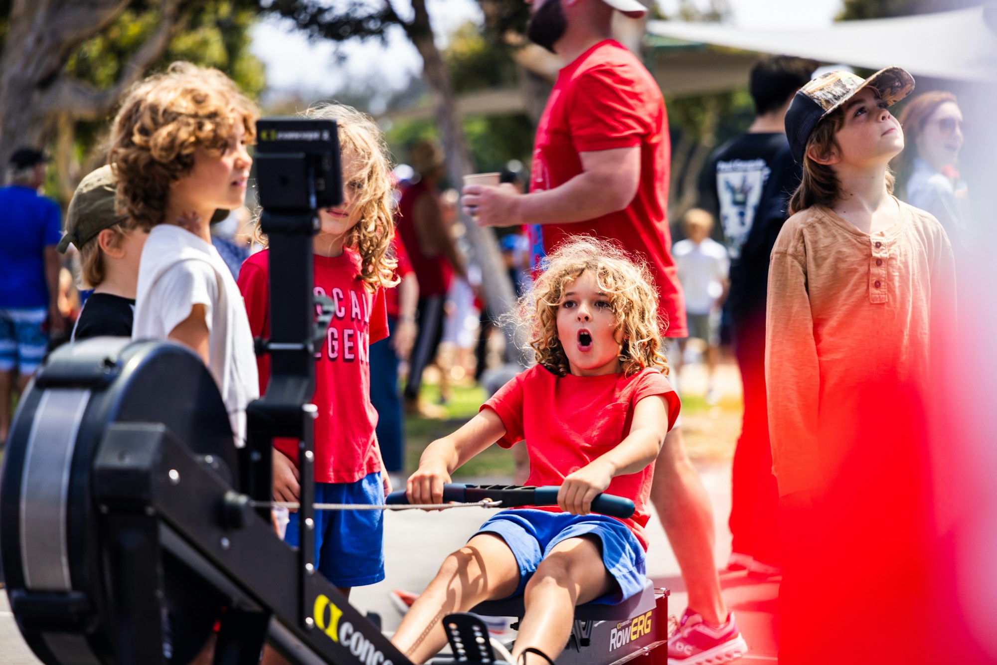 Kids engaging with a rowing machine at an outdoor event, surrounded by onlookers and an adult.