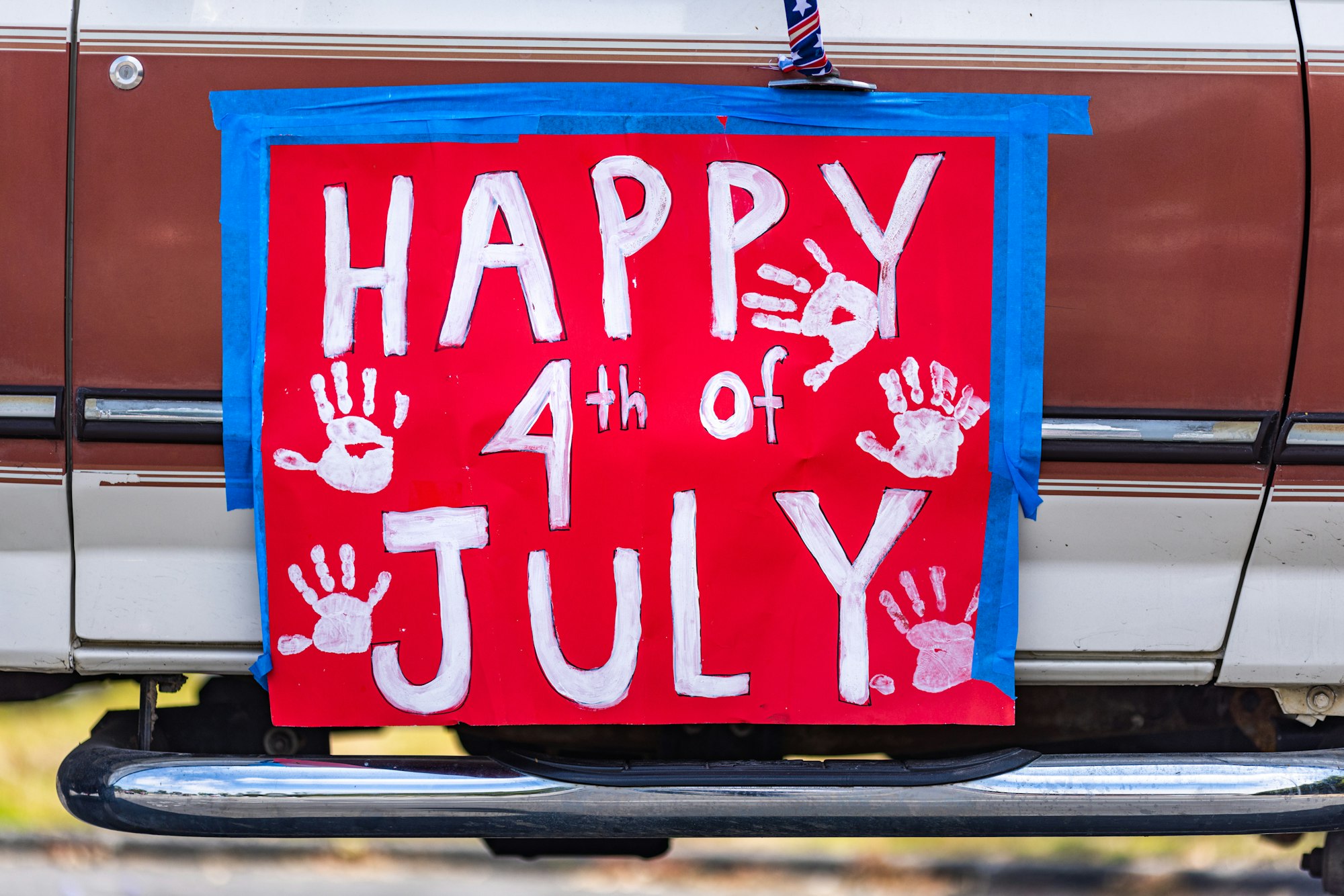 Red sign with "Happy 4th of July" in white letters and handprints, taped to a vehicle.