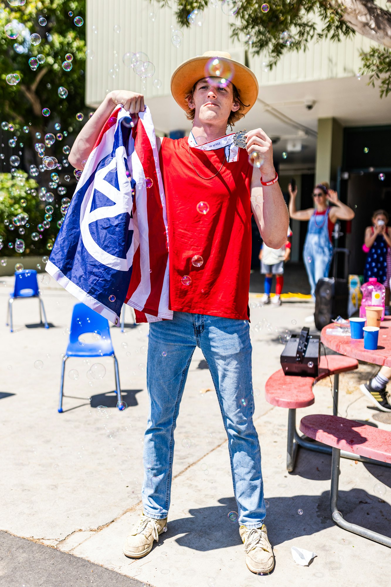 A person in a hat holds a flag and a medal, surrounded by bubbles, with chairs and people in the background.