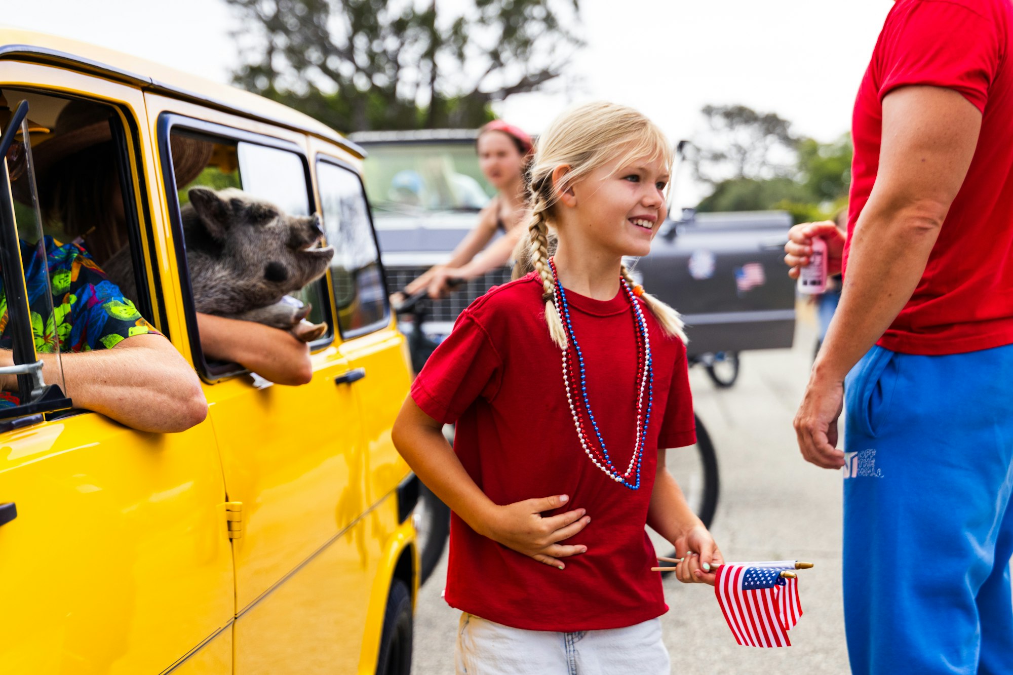 Girl with flag and beads smiling, near a yellow vehicle with a raccoon inside. People are enjoying an outdoor event or parade.