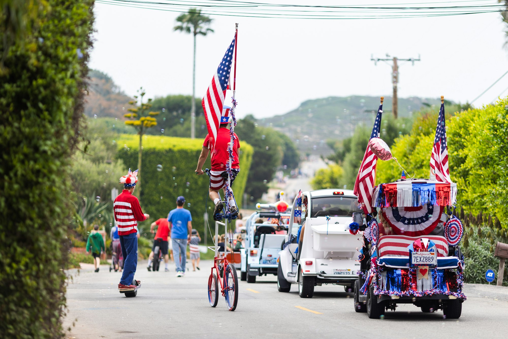 A parade with decorated vehicles, a person on a tall bike, and American flags, celebrating with festive attire.
