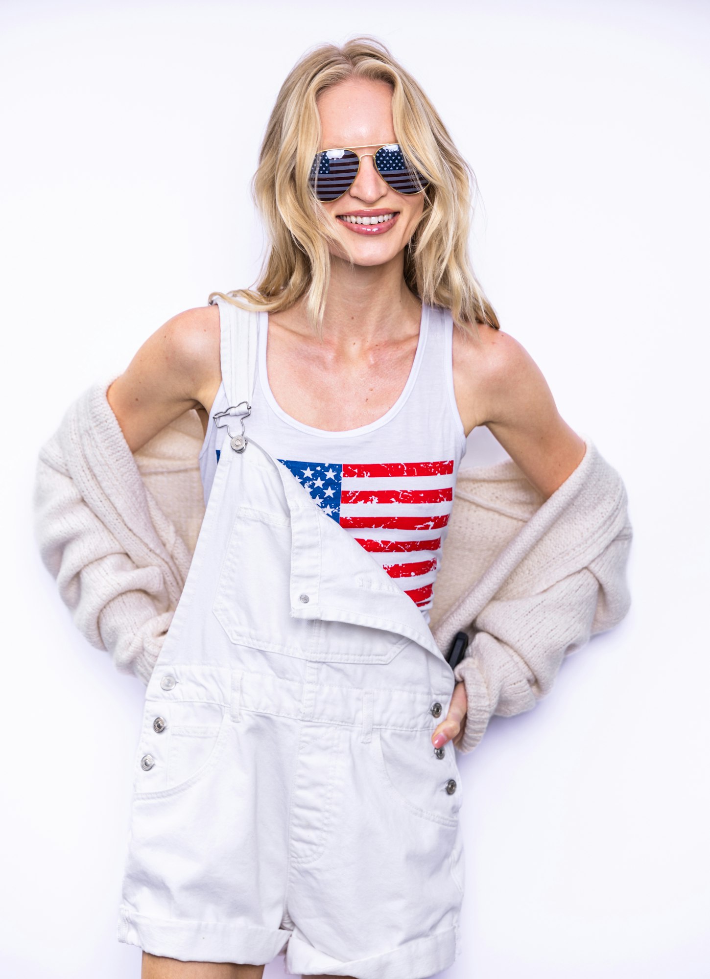 Smiling woman in American flag sunglasses, white overalls, and tank top with flag print, wearing a beige cardigan.