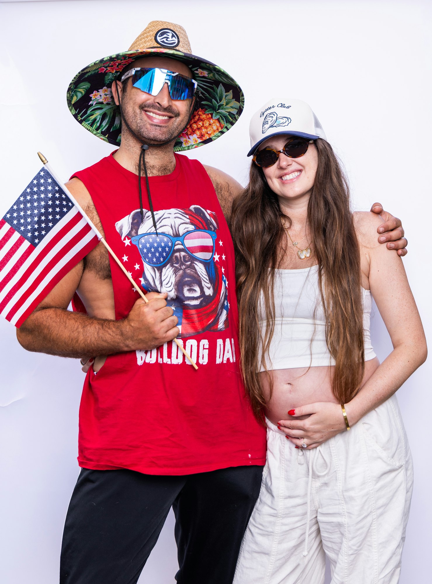 A smiling couple with sunglasses, the man holds a U.S. flag and wears a hat. The woman shows a pregnant belly.