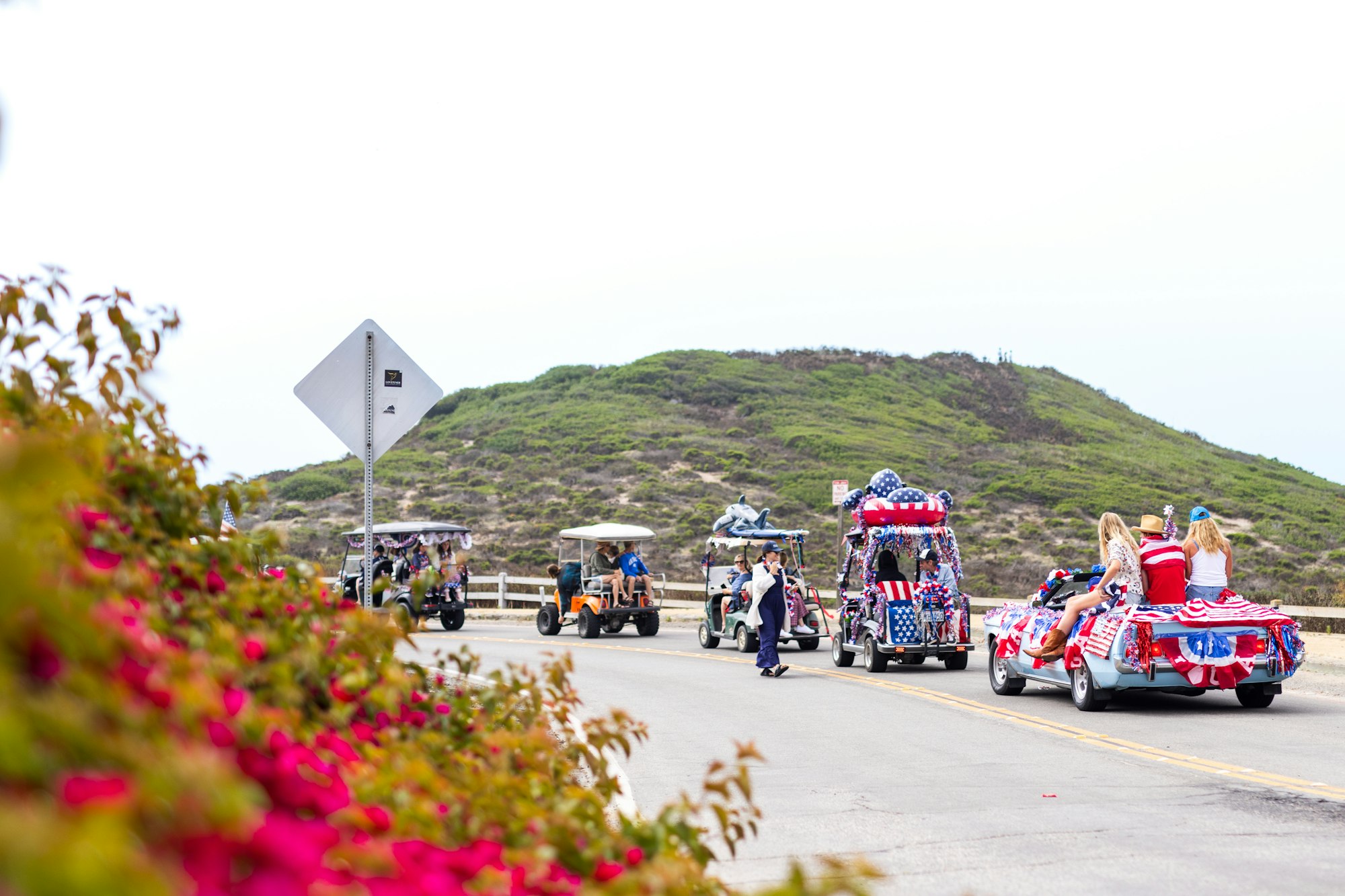 People riding decorated golf carts on a road with a hill in the background and flowers in the foreground.