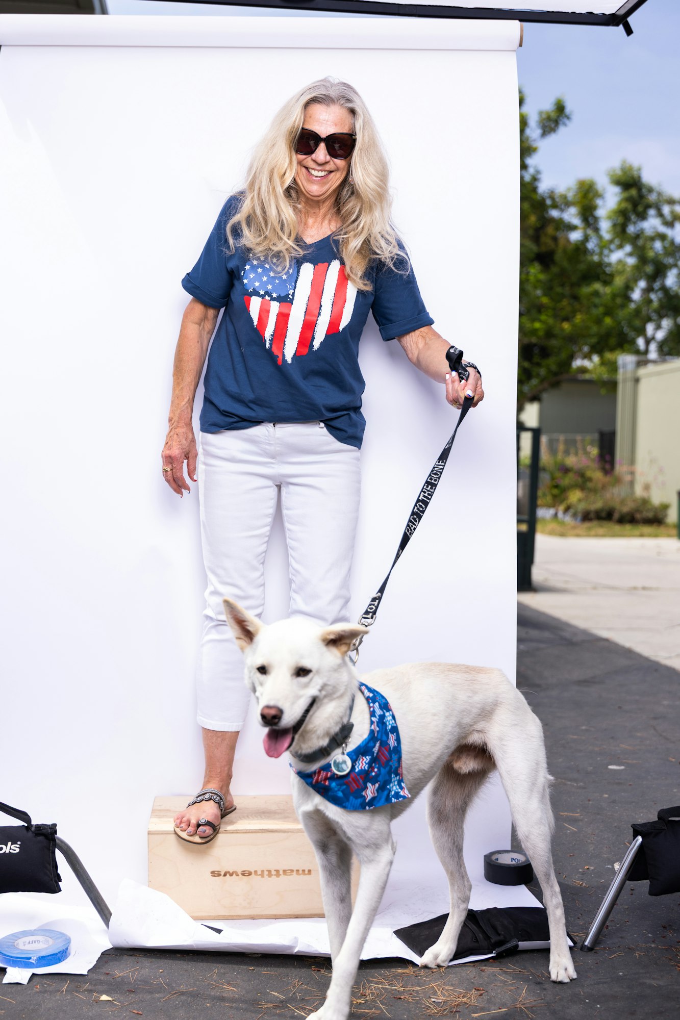 A smiling woman with sunglasses, wearing a flag-themed shirt, stands with a white dog on a leash, in front of a white backdrop.