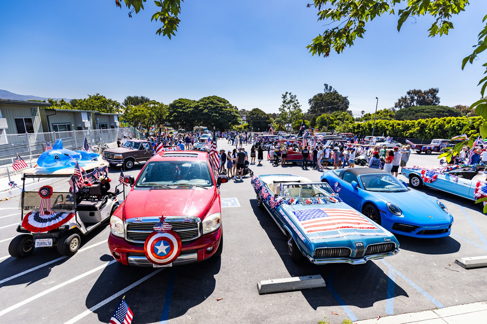 A festive car parade with decorated vehicles, American flags, and people gathering in a parking lot under a clear blue sky.