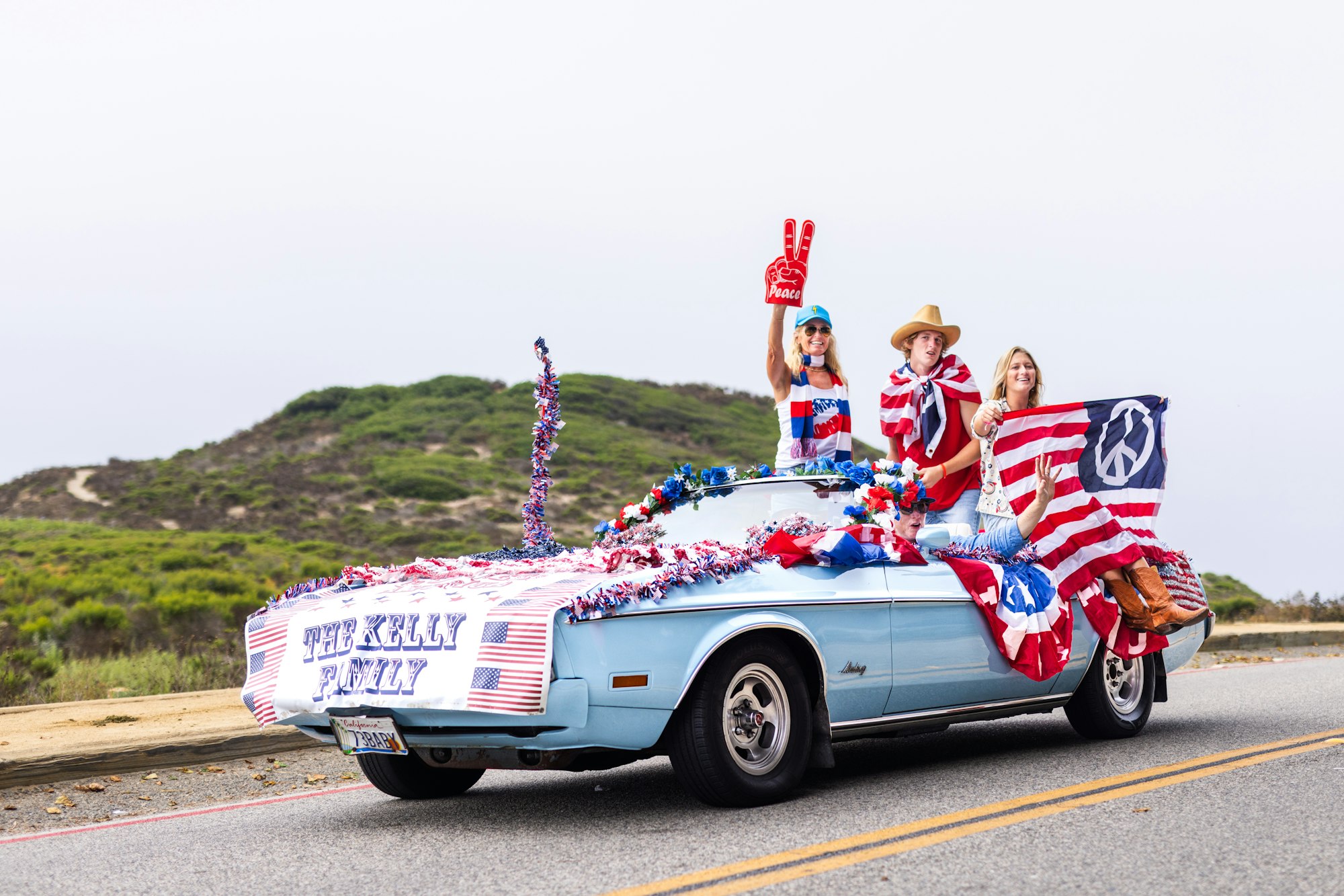 A decorated car with "The Kelly Family," people wearing red, white, and blue, waving flags and a peace sign foam finger.