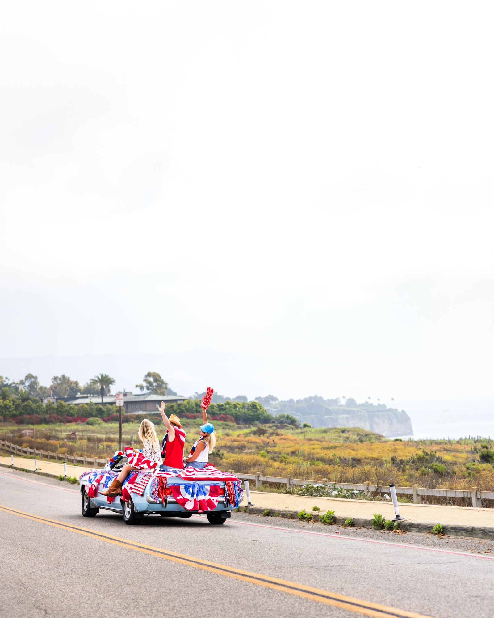 People in a convertible car decorated with red, white, and blue, driving on a coastal road, waving and celebrating.