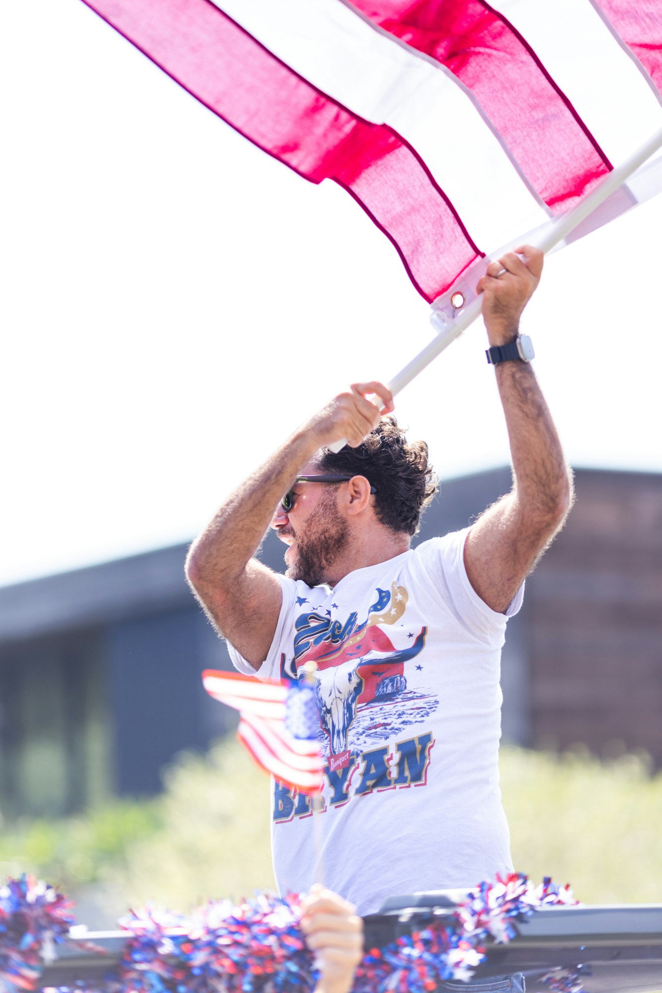A person waving a large red and white flag, wearing sunglasses and a shirt featuring a bull and stars, next to decorated paraphernalia.