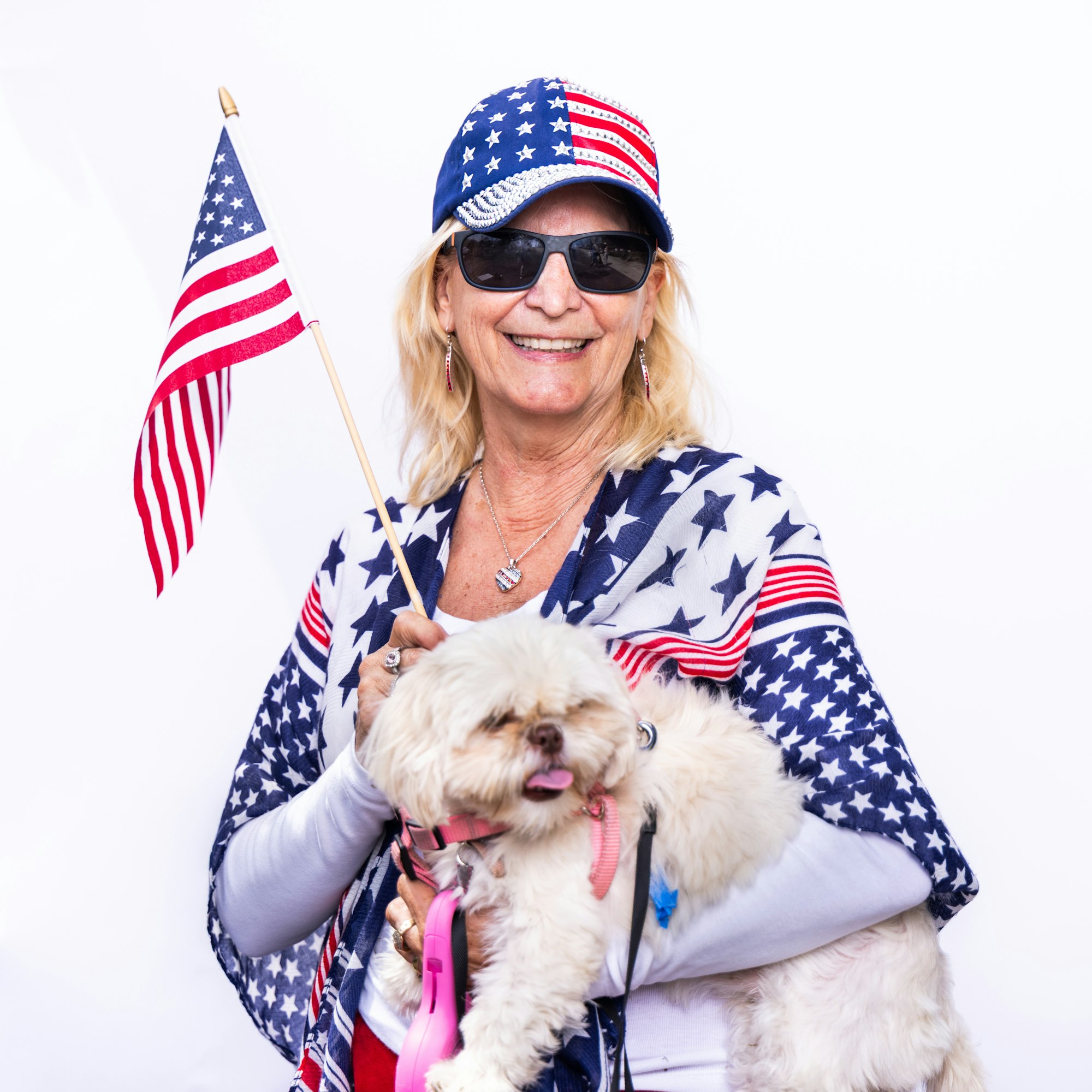 A smiling woman in patriotic attire holds a small dog and an American flag against a white background.