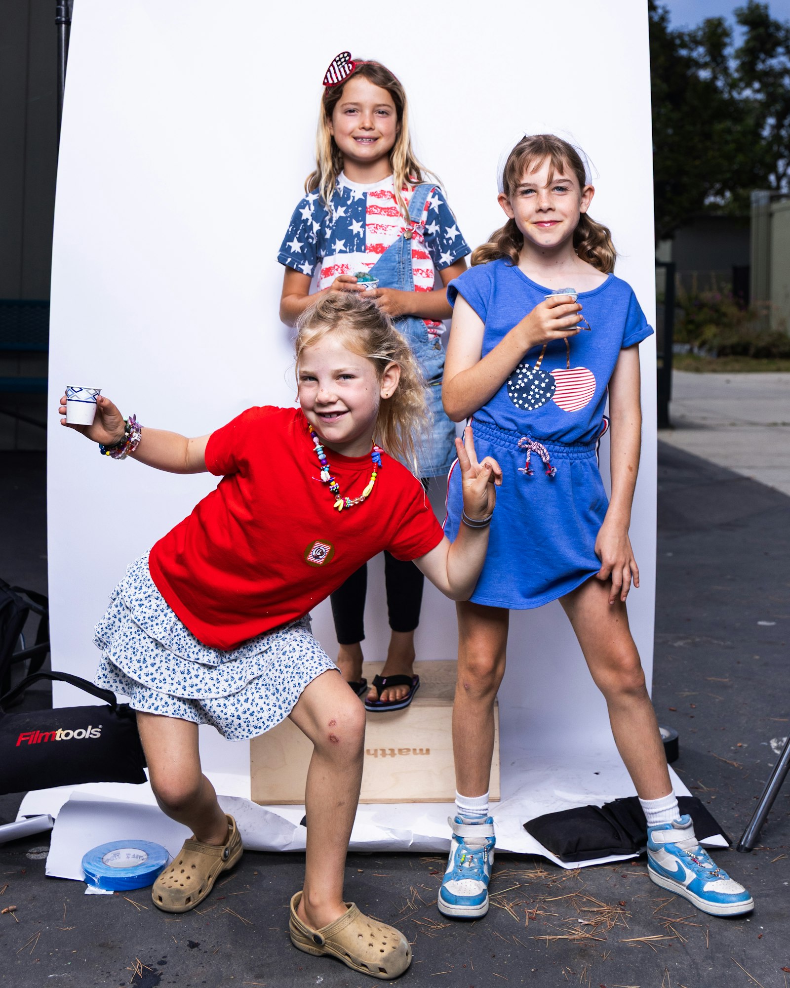 Three kids posing happily in colorful outfits, holding small items, against a white backdrop outdoors.