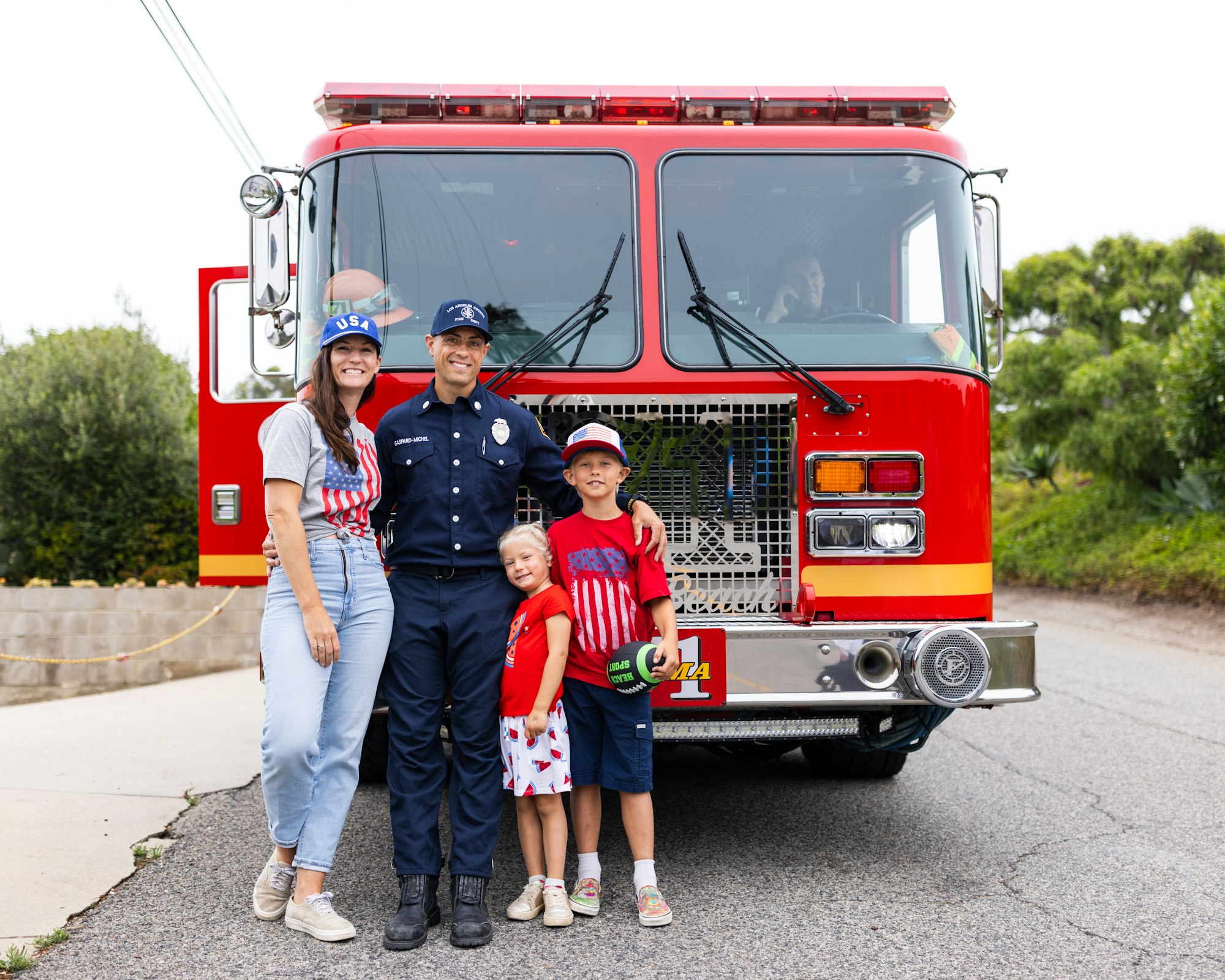 A family poses in front of a fire truck; the firefighter in uniform and others wearing patriotic USA clothing.