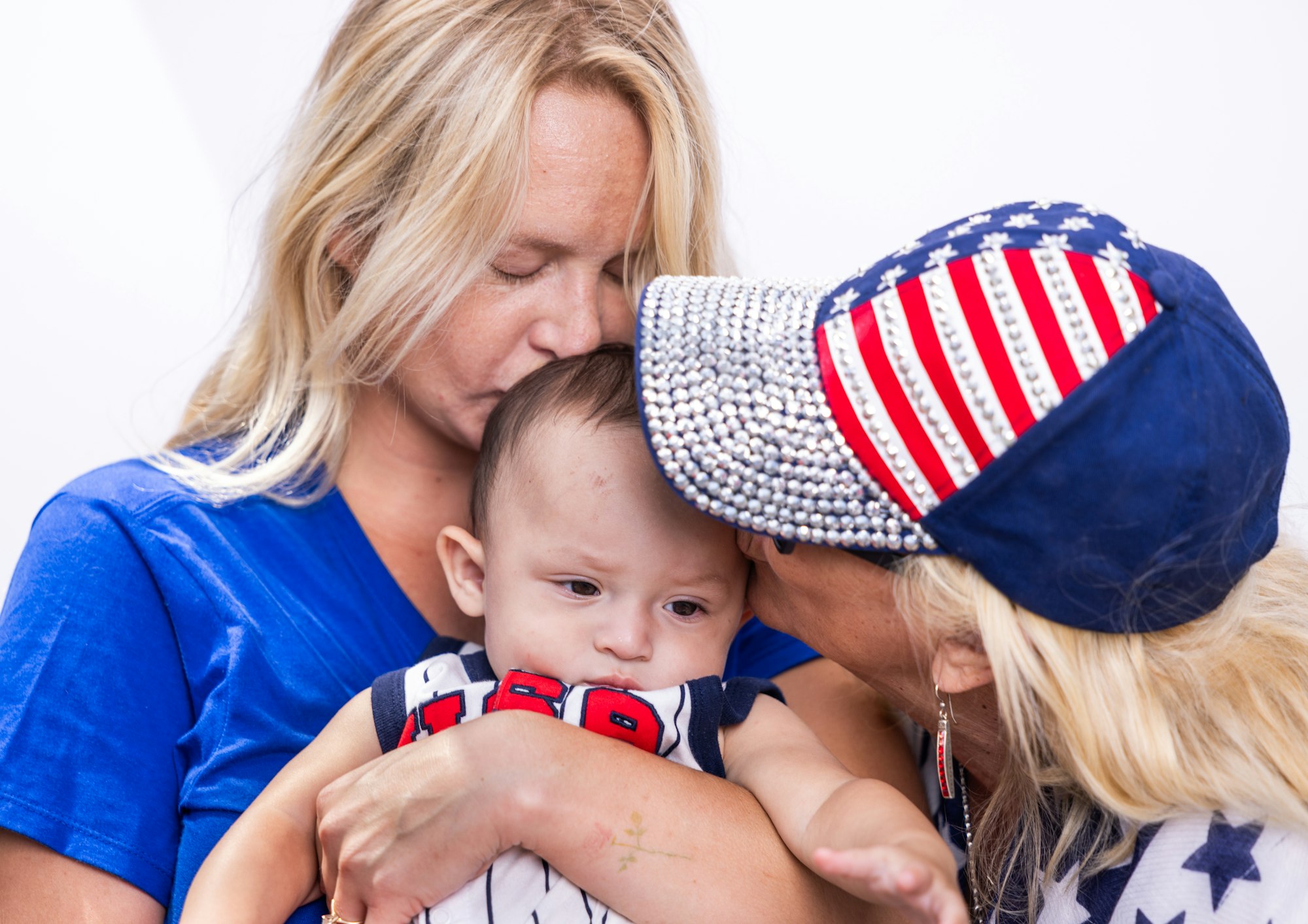 A woman holds a baby while another kisses the baby's head, wearing a patriotic hat with stars and stripes.