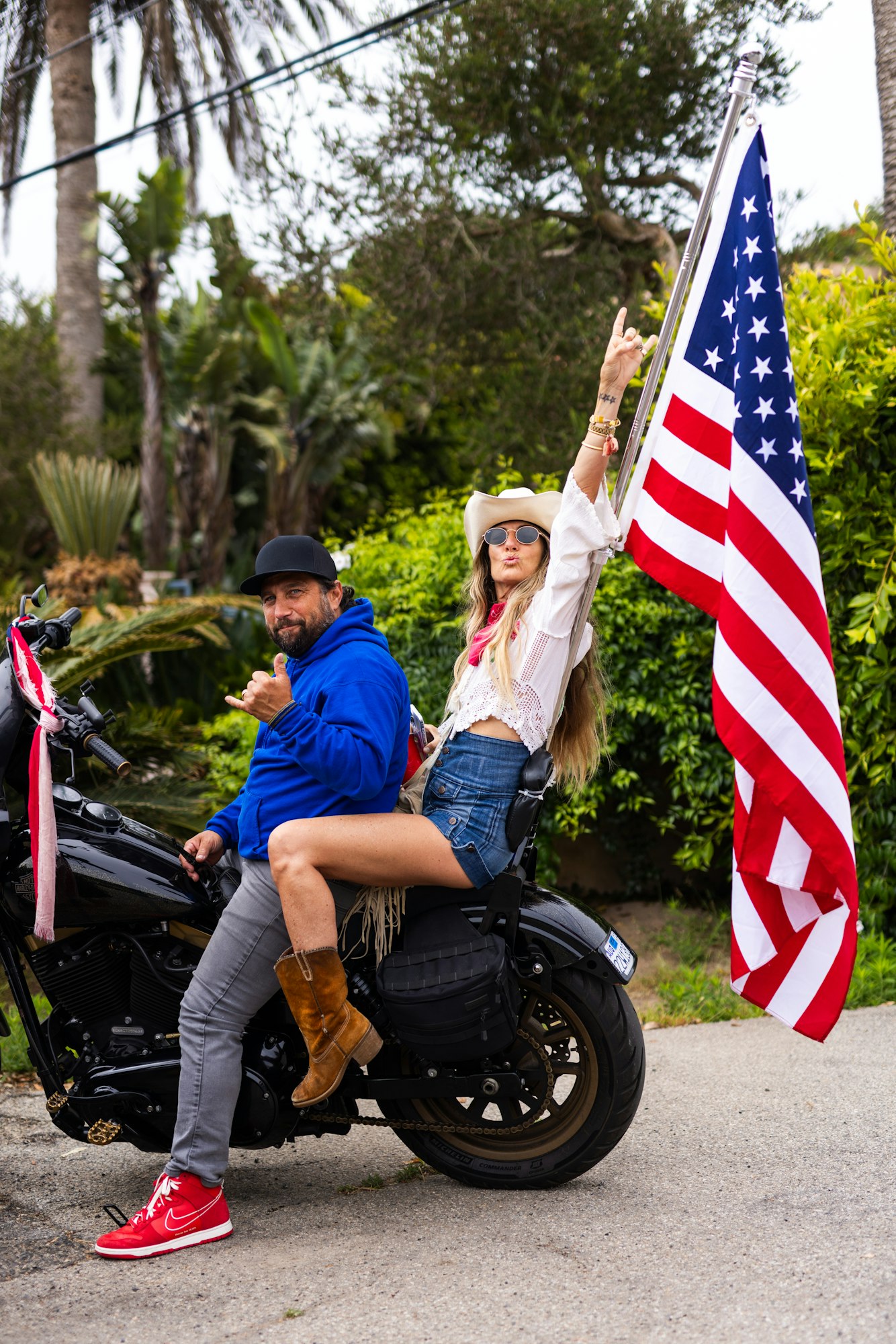 Two people on a motorcycle with an American flag, one giving a peace sign, in a tropical setting.