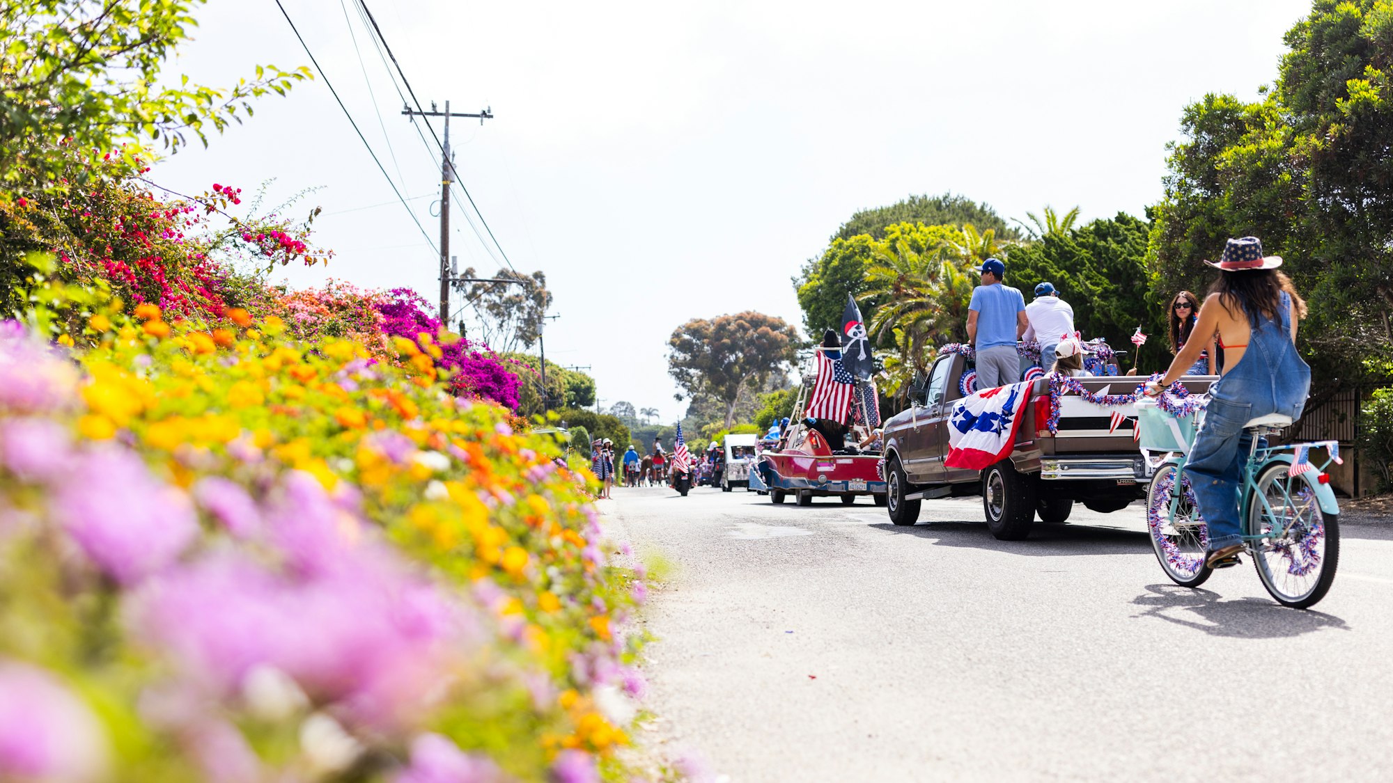 Parade with decorated vehicles and cyclists, American flags, surrounded by colorful flowers and greenery.