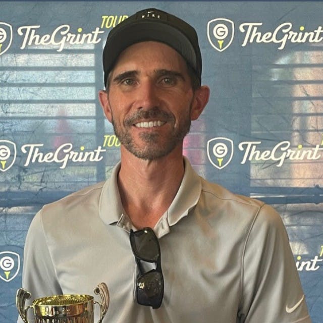 A man wearing a cap, holding a trophy, stands in front of a backdrop with “TheGrint” logos.