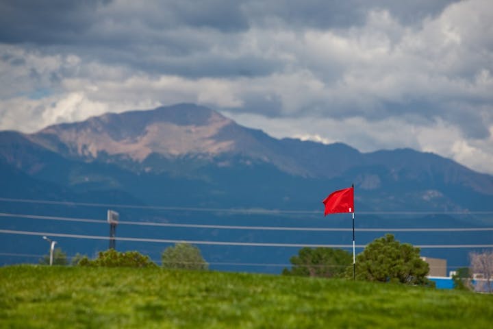 A red flag on a golf course with mountains in the background and dramatic clouds above.