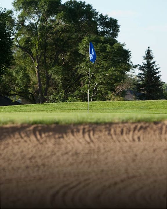 A golf hole with a blue flag and a sand trap in the foreground.
