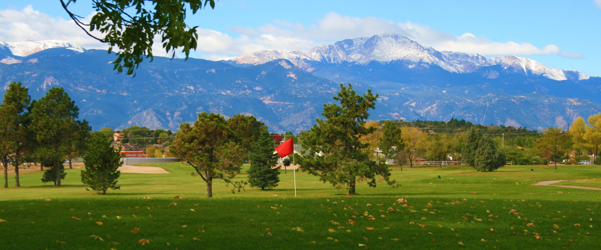 A golf course with trees, a red flag, snow-capped mountains in the background, and a clear sky.