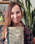 A smiling person holding a book, with plants and bookshelves in the background.