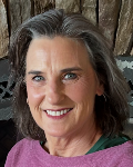 A smiling woman with gray hair, wearing a pink sweater, is posed in front of a wooden background.