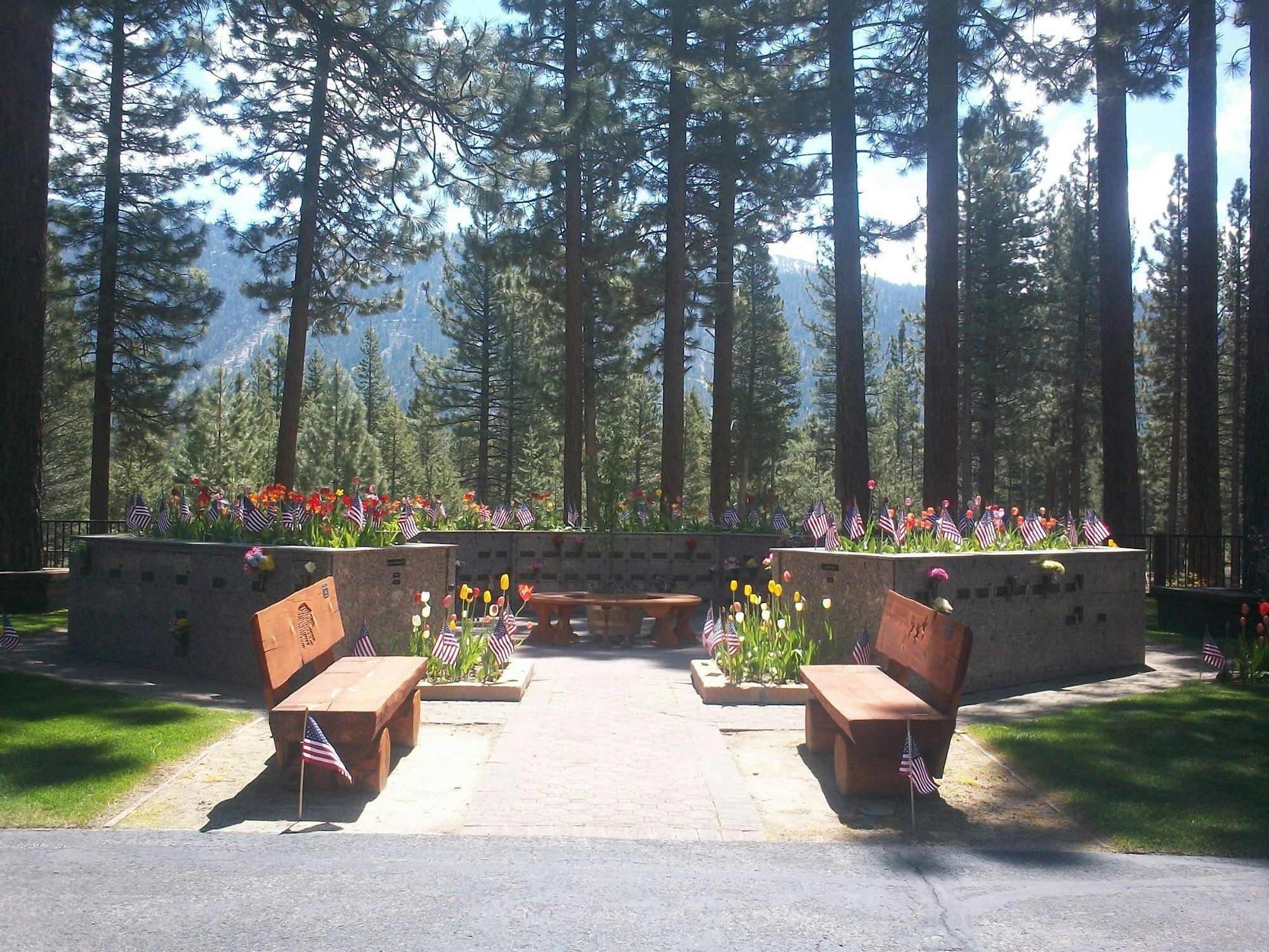 A serene memorial garden with flowers and American flags, surrounded by tall trees and mountains in the background.
