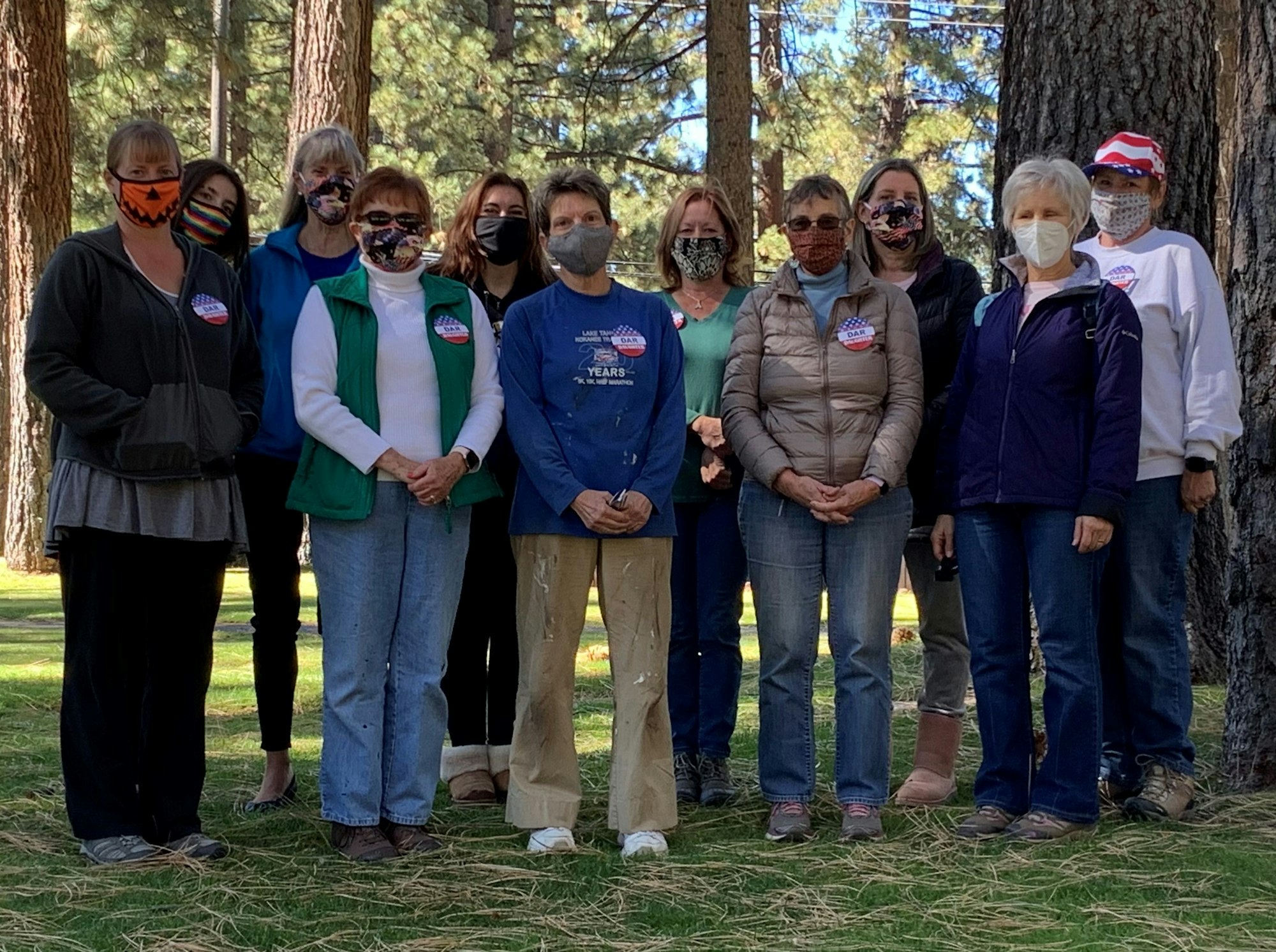 A group of women wearing masks poses together outdoors, surrounded by trees, some showing buttons or stickers.