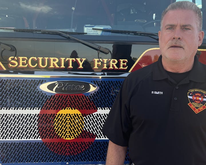 A man in a black fire department uniform stands in front of a fire truck displaying "Security Fire" and a Colorado emblem.