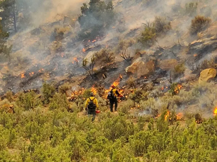 Firefighters are battling a wildfire, surrounded by flames and smoke in a rugged, hilly terrain.