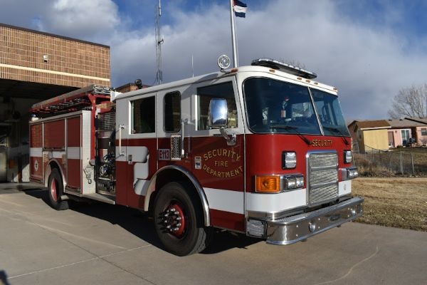 A red and white fire truck from the Security Fire Department, parked outside a fire station with a cloudy sky.