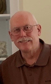 The image shows a smiling older man with glasses and a mustache, wearing a brown polo shirt, set in a light interior.