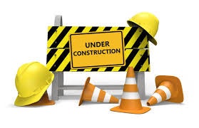 A sign saying "UNDER CONSTRUCTION" with a hard hat, traffic cones, and caution barrier.