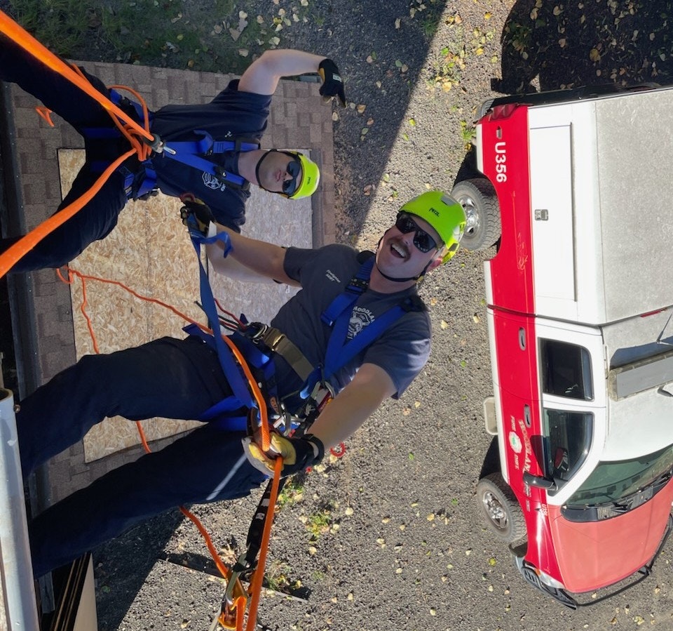 Two people in safety gear are rappelling down, smiling, with a red vehicle in the background.