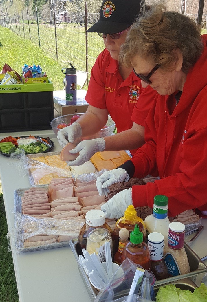 Two people in red shirts preparing food with condiments and snacks on a table.