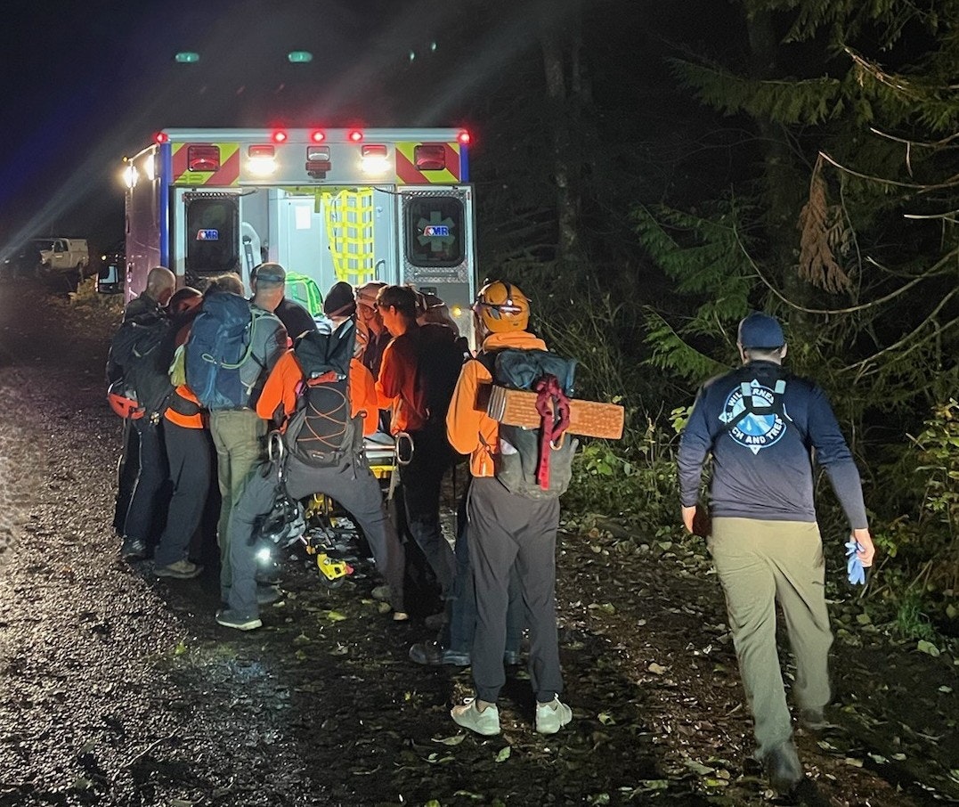 Rescue team members in bright orange shirts gather around an ambulance at night, assisting with an emergency situation in the woods.