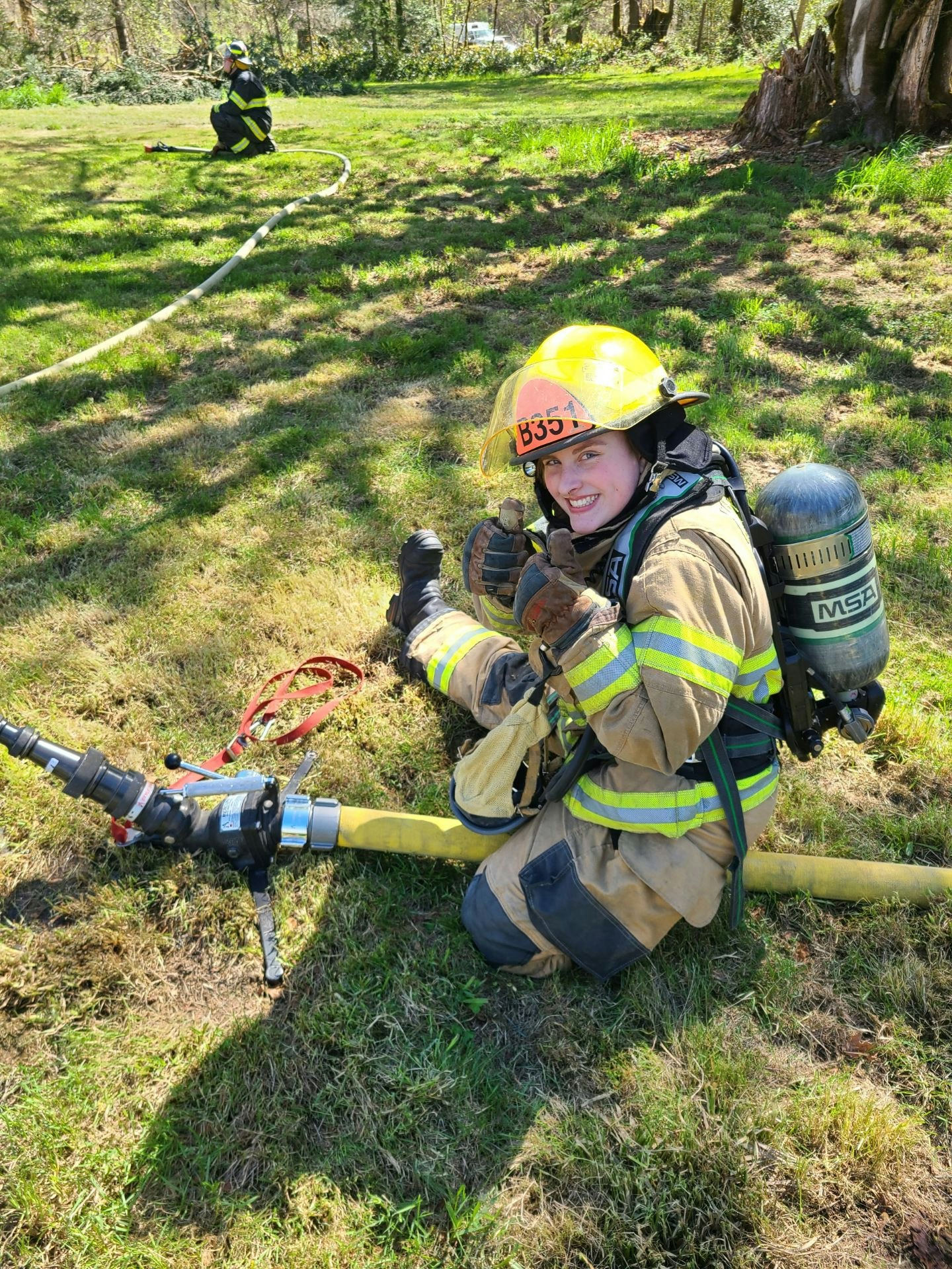 Two firefighters in gear sitting on grass, one smiling at the camera, with hoses and equipment.