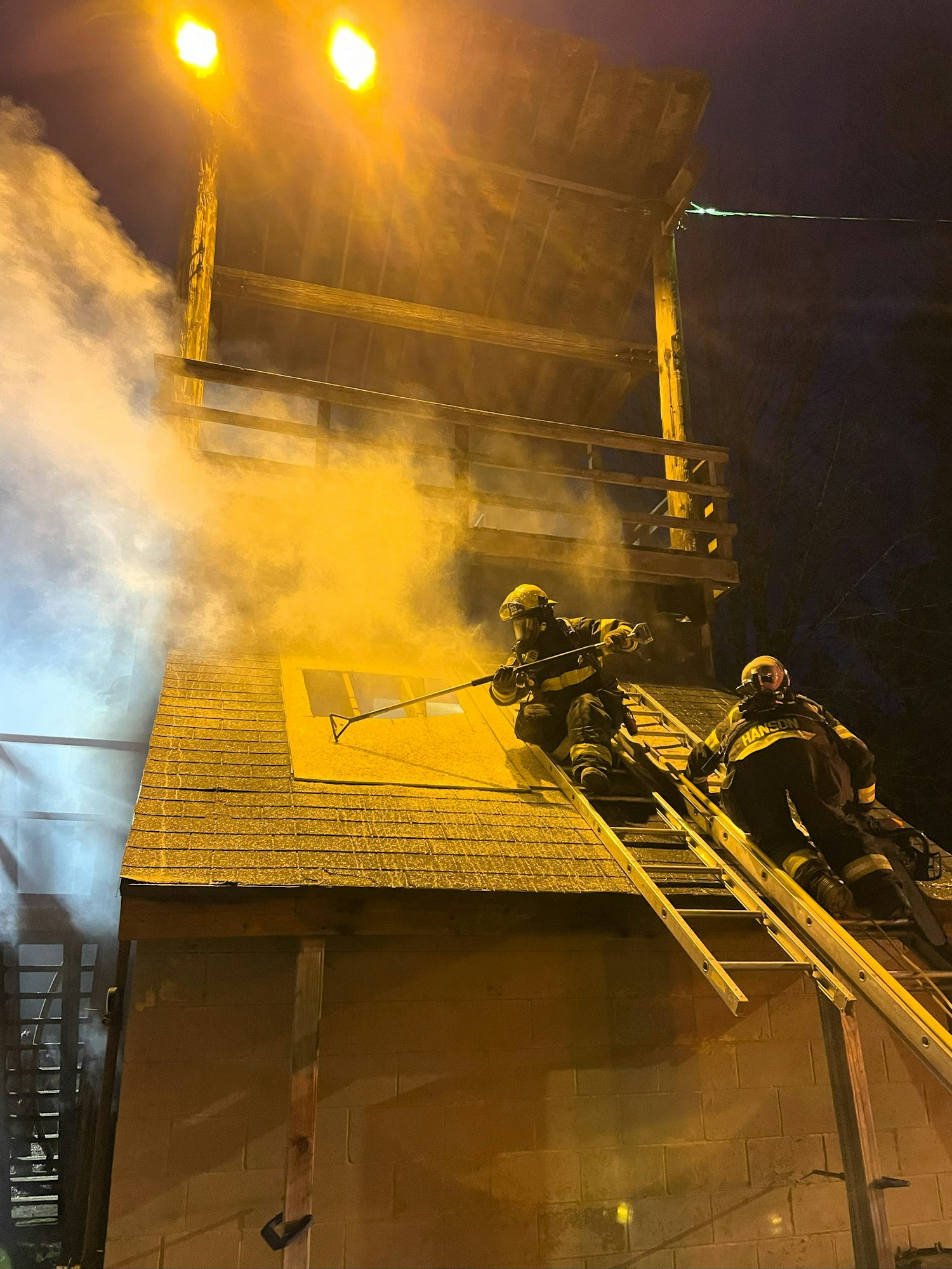 Firefighters on a ladder, smoke billowing from a building during night.