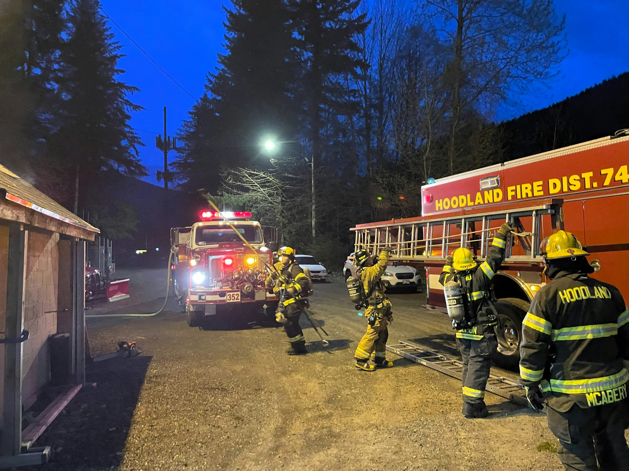 Firefighters in gear stand near fire trucks, one with a hose, another with a ladder. It's a nighttime scene with trees and streetlights.