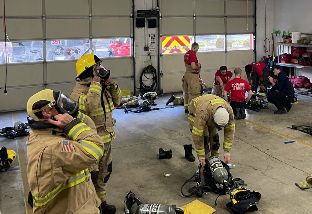 Firefighters in gear prepare equipment in a fire station. Some are putting on masks while others organize gear.