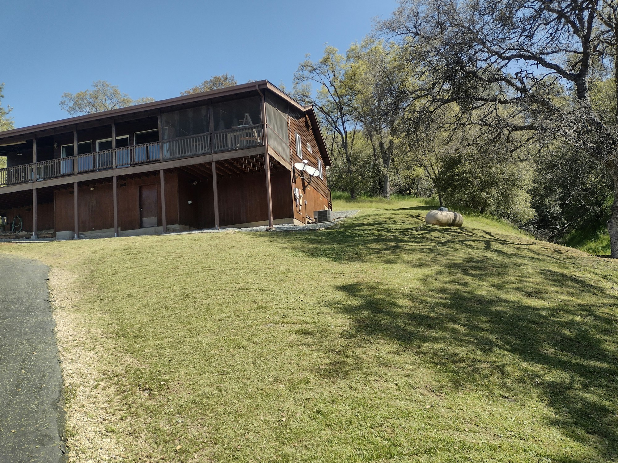A wooden house on a sloped grassy hill, surrounded by trees under a clear blue sky.