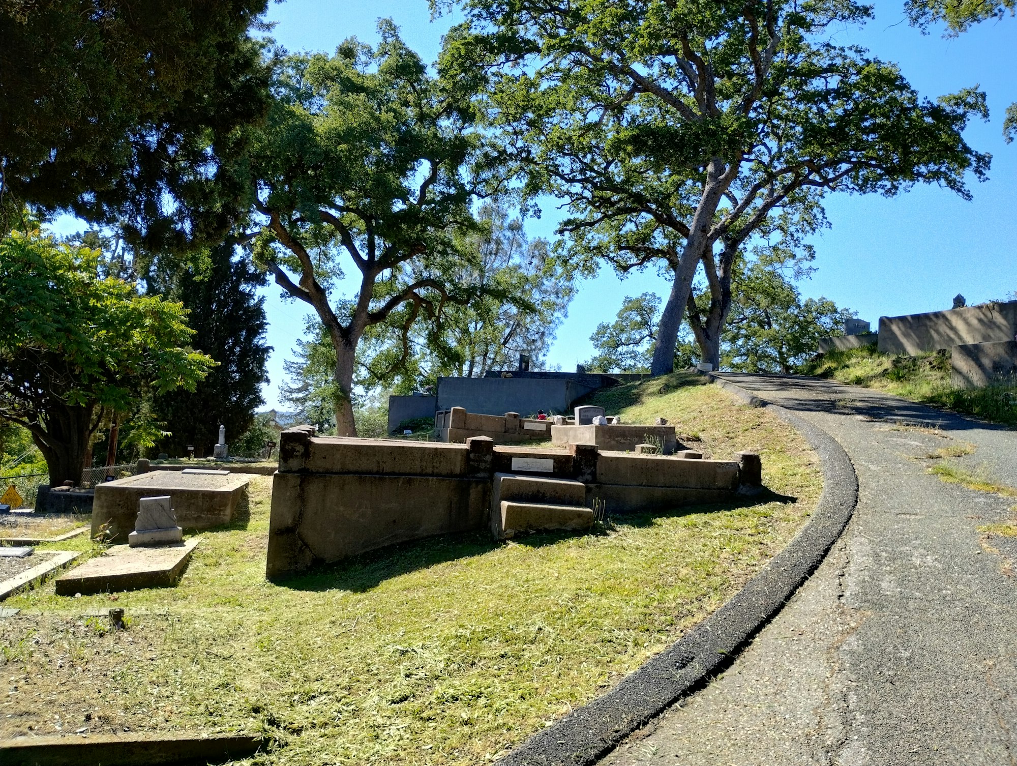 A cemetery with gravestones on a grassy hill, surrounded by trees under a clear blue sky.