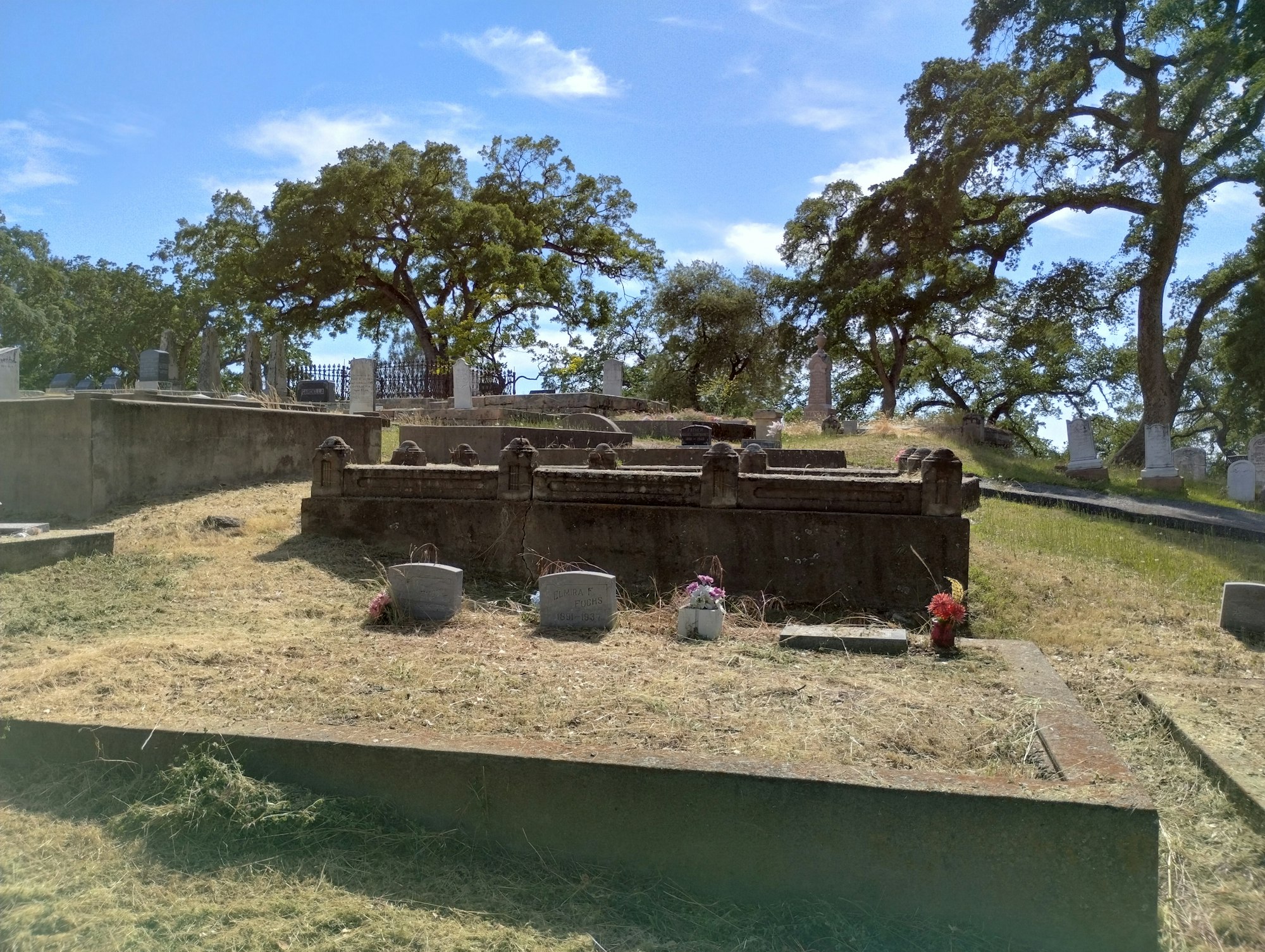 A cemetery with gravestones, trees, grass, and a clear blue sky.