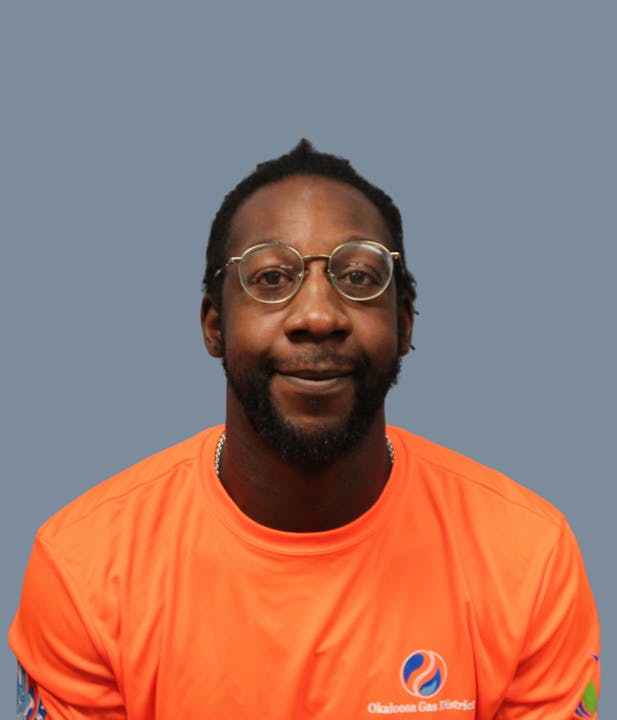 The image shows a man with glasses wearing an orange shirt, smiling against a plain gray background.