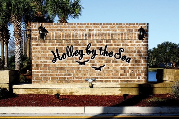 A brick sign reading "Holley by the Sea" with birds, surrounded by palm trees and landscape.