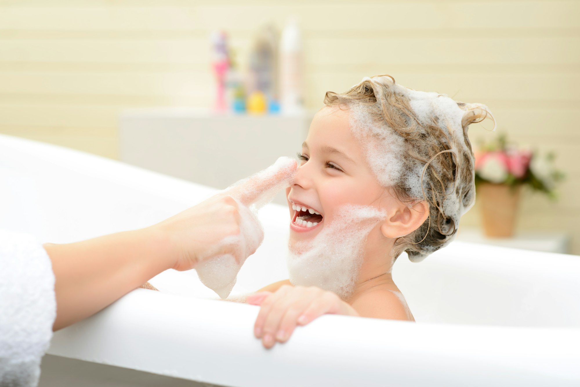 A child with soap bubbles laughs in a bathtub as someone playfully touches their nose.