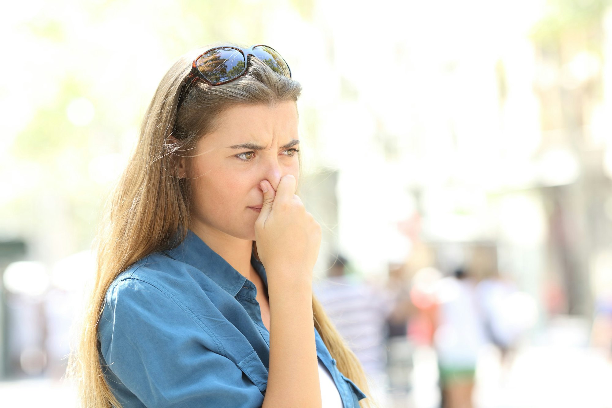 A woman outdoors pinching her nose, showing discomfort, wearing sunglasses on her head.