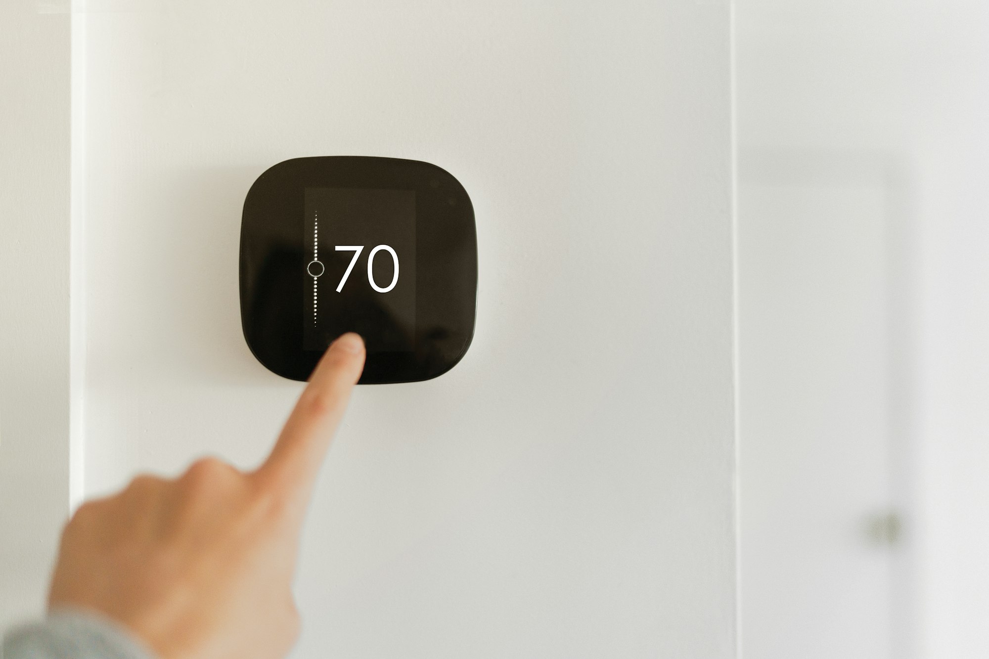 A finger is adjusting a digital thermostat set to 70 degrees on a wall.
